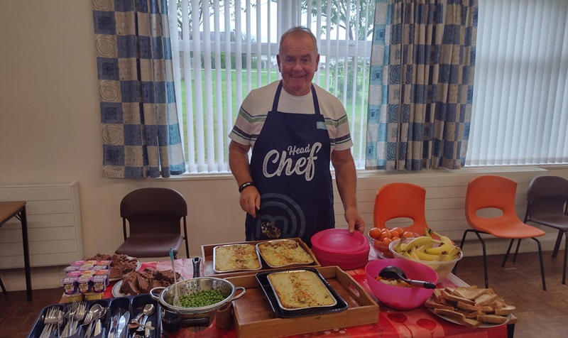 A man standing behind a table that has foot, plates, dishes. He is wearing a blue apron with the word 'chef'