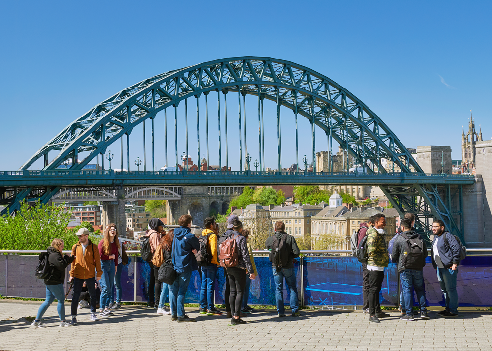 The Tyne Bridge in Newcastle with a group of people stood on a pavement looking into the distance at the bridge