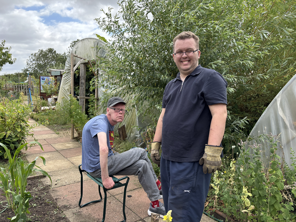 Two people outside in an allotment, one is standing with gloves on, the other is sat down