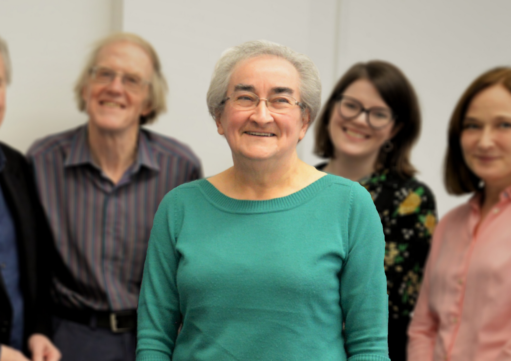 Image of Rosemary who is wearing glasses and a green jumper. In the background is three people who are slightly blurred to focus on Rosemary