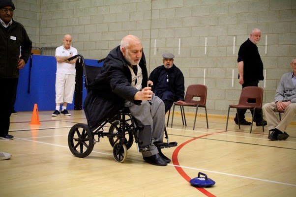 A man sitting in a wheelchair on an indoor court. He has just let go of a curling stone with his team watching on