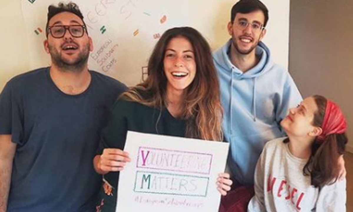 Four people looking at the camera with the person in the middle holding a handmade sign saying Volunteering Matters