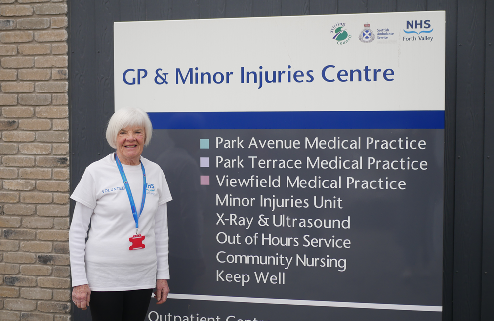 Older person in a white NHS tshirt standing next to an NHS Forth Valley sign which says GP and minor injuries centre