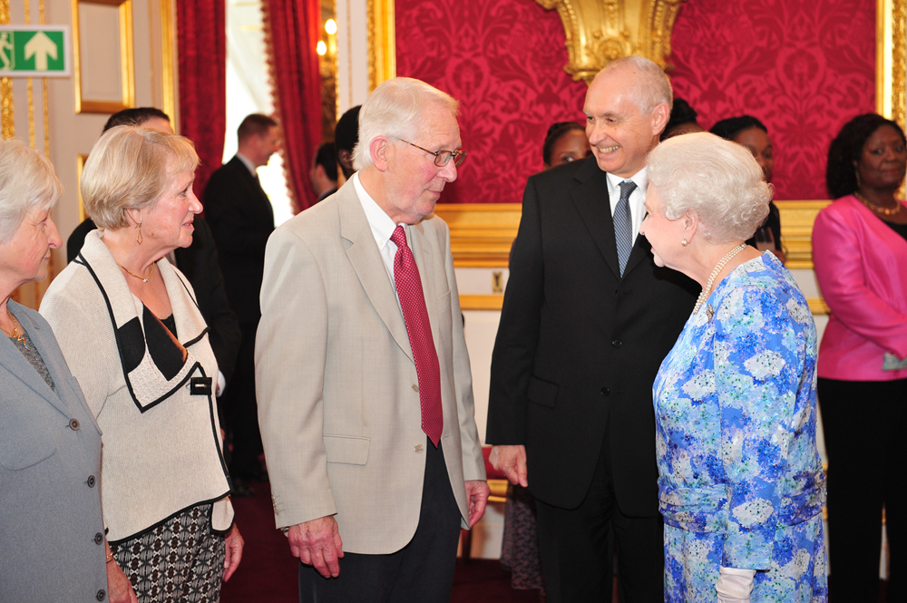 Four individuals, Colin standing in a black jacket, speaking to the late HM Queen Elizabeth inside a room with red and golden wallpaper