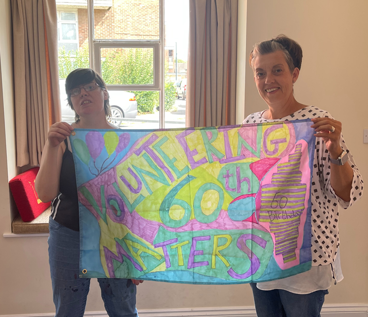 Helen with another person holding up a fabric banner with Volunteering Matters 60th stitched into it