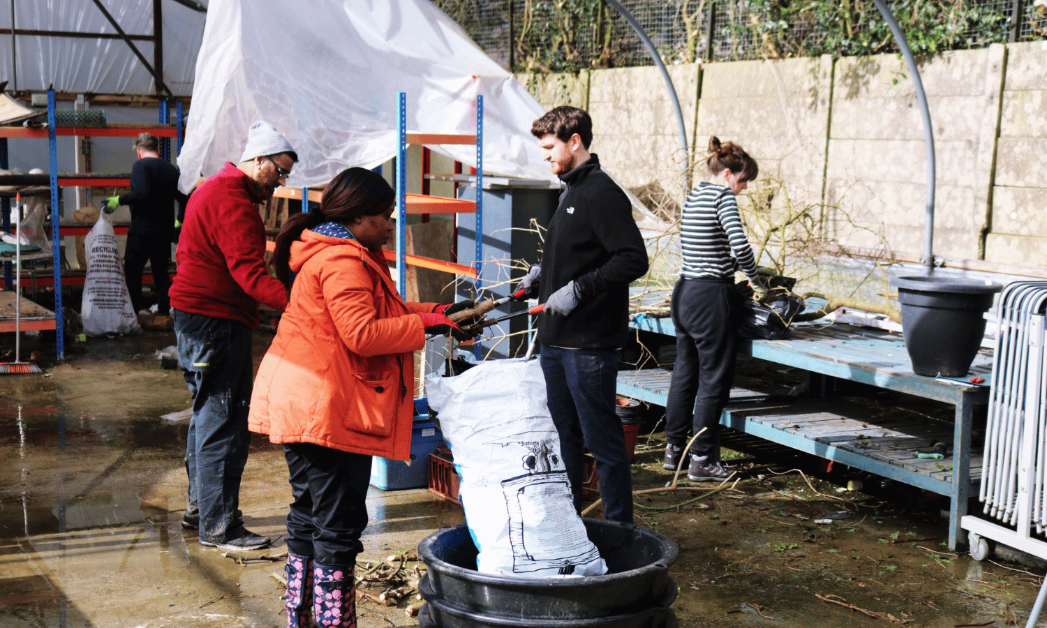 A woman putting items in a white bag outside, wearing a red outdoor jacket. There are four colleagues in the background doing various tasks