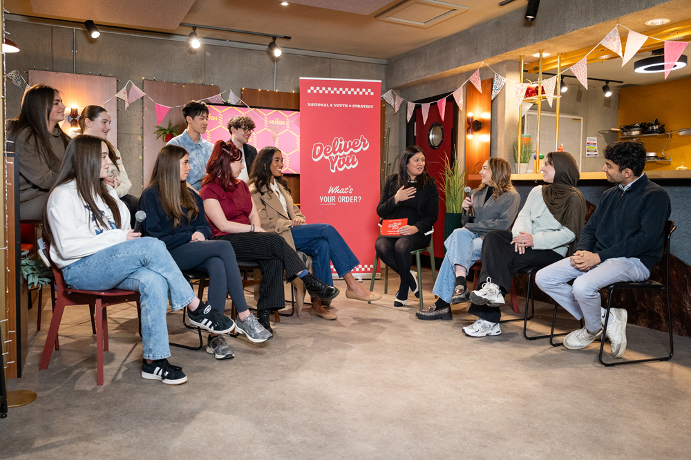 Young people sitting next to Lisa Nandy MP with a red banner with Deliver You on